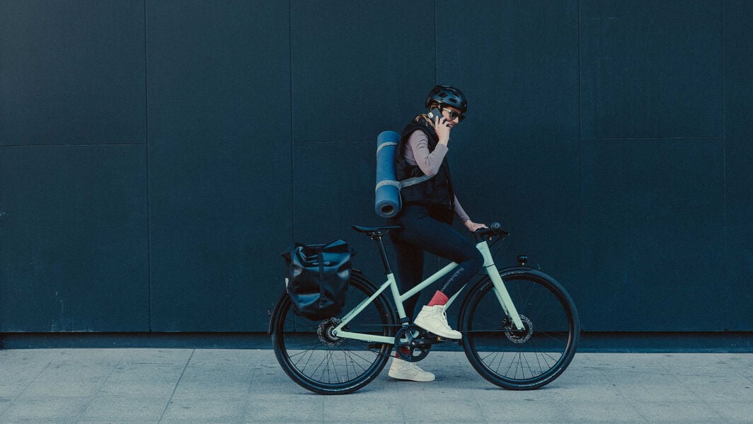 Explore the city in comfort with the perfect city bike — designed for smooth, everyday rides Man sitting on stone steps beside a Canyon bike, adjusting his camera in a park setting.